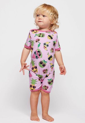 Kleinkind mit lockigem blondem Haar, das einen pinkfarbenen Overall mit bunten, Sonnenbrillen tragenden Obstcharakteren trägt, barfuß vor einem einfarbigen Hintergrund stehend.