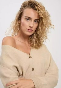 Beżowy sweter z dzianiny o dekolcie off-the-shoulder, z dużymi guzikami i fakturowanym materiałem. Miękki, luźny krój.