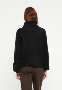 Un pull noir surdimensionné avec un col haut, de larges manches avec des poignets repliés et une texture travaillée. Portez-le sur un pantalon marron.