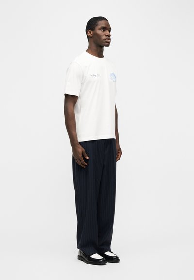 Άνδρας που στέκεται, φοράει λευκό κοντομάνικο μπλουζάκι με μπλε γραφικό, σκούρο παντελόνι με λεπτές κάθετες ρίγες και φαρδιά μπατζάκια, και μαύρα και λευκά επίσημα παπούτσια, ανοιχτό φόντο.