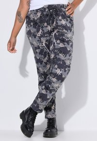 Grafitgrå joggerbukser med camouflageprint, der har snøre i taljen, lommer og forskellige tekstdesigns. Båret med sorte snørebåndsstøvler.