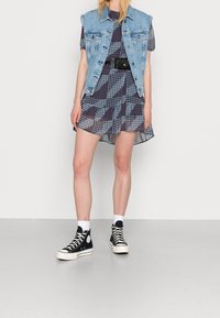 Jeansweste über einem gemusterten marineblauen Kleid mit gerafftem Saum, kombiniert mit schwarzen High-Top-Sneakers und weißen Knöchelsocken.