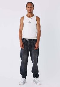 Canottiera bianca con logo nero, jeans scuri, cintura nera e sneaker bianche con dettagli grigi. Modello in piedi contro uno sfondo neutro.