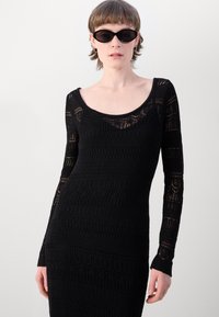 Abito nero a maglia con maniche lunghe, caratterizzato da un motivo testurizzato e scollatura rotonda. Modella che indossa occhiali da sole neri slim.