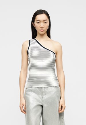 Young woman with long dark hair wearing a one-shoulder white top with thin black stripes and light gray pants, standing against a plain background.