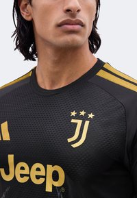 Gros plan d'une personne portant un maillot de football noir avec le logo doré de la Juventus, trois étoiles, des bandes dorées sur les épaules et le logo du sponsor Jeep.