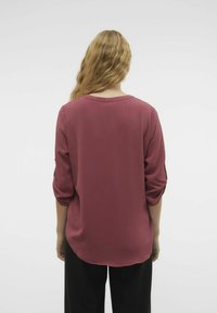 Blouse bordeaux avec une texture fluide, encolure ronde et manches trois-quarts. Tissu lisse avec un ourlet arrondi, associé à un pantalon noir.