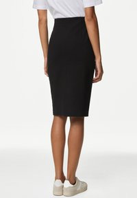 Schwarzer Bleistiftrock mit hohem Bund, figurbetonter Silhouette und glatter Textur, kombiniert mit hellen Sneakers, von hinten gesehen.