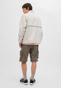 Lichtbeige windjack met een hoge kraag en zwarte bies, gecombineerd met bruine cargoshorts met zijzakken, zwarte sneakers en grijze sokken.