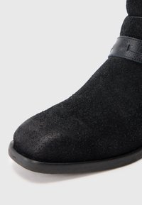 Černá semišová kotníková bota s kulatou špičkou, gumovou podrážkou a koženým páskem kolem horní části.