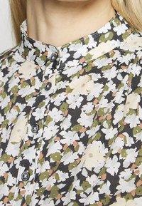 Bloes met bloemenprint op een zwarte achtergrond, met witte, groene en perzikkleurige bloemen. Inclusief een kraag en knoopsluiting bij de hals.