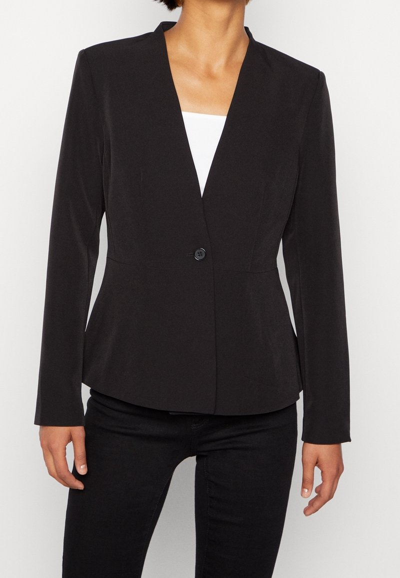 Femme portant un blazer noir cintré avec un bouton sur un haut blanc et un pantalon noir sur un fond clair uni.