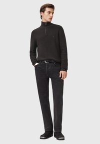 Man met een zwarte ritsvest, zwarte jeans, een zwarte riem en zwarte laarzen, staand met één hand in de zak tegen een effen achtergrond.