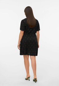 Robe noire à manches courtes et taille cintrée, ornée d'un motif de cerises. Associée à des chaussures à talons verts, vue de dos.