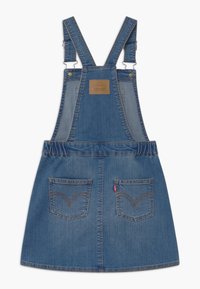 Džínová zástěra s nastavitelnými ramínky, elastickým pasem, dvěma zadními kapsami a hnědou logo nášivkou nahoře. Modrá barva, ležérní design.