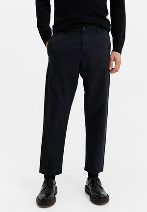 Mann trägt schwarze knöchellange Hosen, schwarzes Langarmshirt, schwarze Socken und glänzende schwarze Schnürschuhe vor einfarbigem Hintergrund.