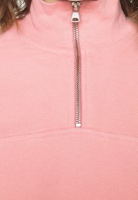 Gros plan d'un pull rose avec une demi-fermeture éclair argentée et un col montant côtelé visible sur le cou d'une personne.