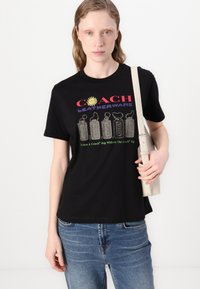 Camiseta de algodón negra con gráfico de "COACH LEATHERWARE" en rojo y púrpura, que presenta diseños de botellas coloridas y texto abajo. El modelo sostiene una bolsa clara.