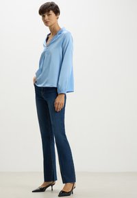 Blusa azzurra chiaro a maniche lunghe con colletto, abbinata a jeans blu scuro e décolleté neri a punta, caratterizzati da gamba dritta e orlo sfilacciato.