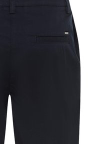 Primer plano de pantalones azul marino oscuro que muestran el bolsillo trasero con una pequeña etiqueta metálica de la marca en el lado derecho.