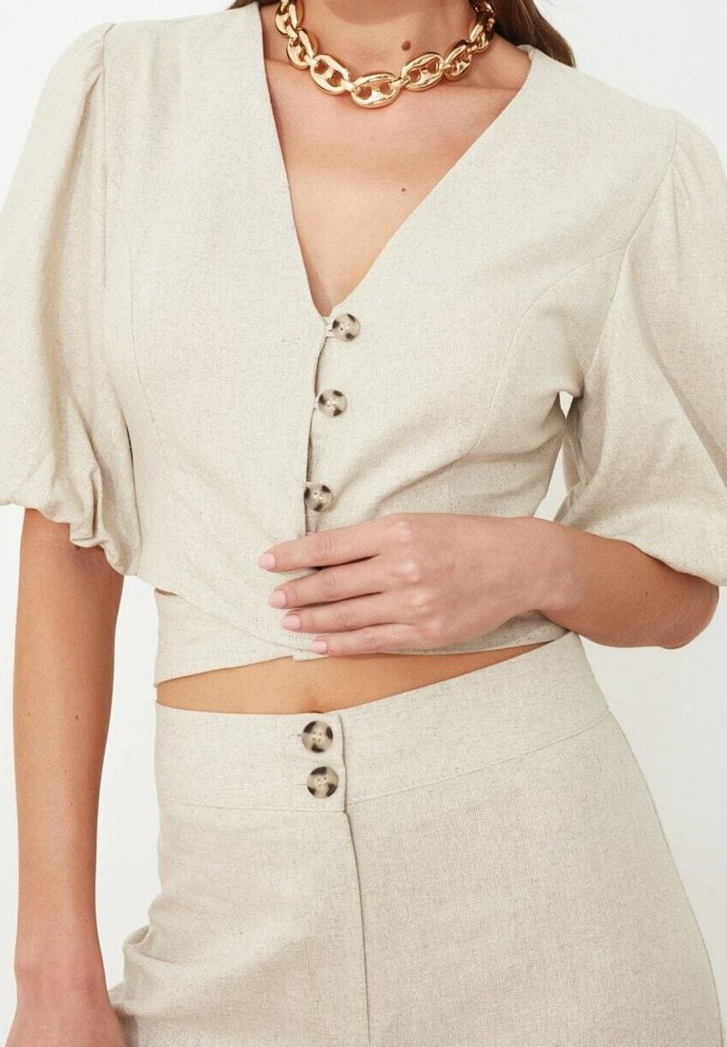 Femme portant un top court beige à manches bouffantes et une jupe taille haute assortie, accessoirisée d'un collier chaîne dorée épaisse.