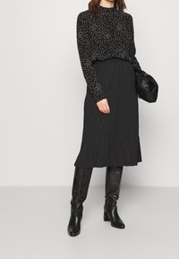 Zwarte blouse met lange mouwen en witte polka dots, gecombineerd met een zwarte plissérok en kniehoge zwarte leren laarzen, met een zwarte handtas in de hand.