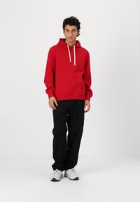 Rød hettegenser med frontlomme og hvite snorer, kombinert med svarte bukser og hvite sneakers. Enkelt design, avslappet passform, myk stoff.