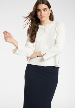 Eine Frau mit langen braunen Haaren trägt einen weißen, geknöpften Pullover mit weiten Ärmeln und einen dunkelblauen Rock und posiert vor einem schlichten Hintergrund.