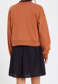 Sweatshirt marron court avec un col rond et des épaules tombantes, associé à une jupe noire plissée. Le tissu semble doux et décontracté.
