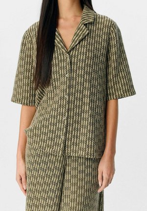 Vrouw draagt een olijfgroen-wit shirt met korte mouwen en zigzagpatroon met bijpassende rok, staand tegen een effen achtergrond.