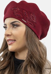 Béret en tricot bordeaux avec un motif texturé, des accents en cristaux et une étiquette de marque en argent sur le côté. Matériau doux et confortable.