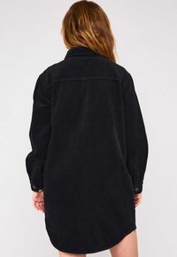 Abito camicia in velluto a coste nero con colletto, maniche lunghe e polsini abbottonati. Presenta una superficie testurizzata e un orlo asimmetrico.