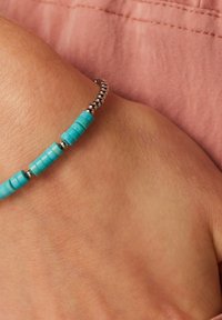 Turfroze kralenarmband met cilindrische kralen en zilveren metalen accenten, gedragen om een lichtgekleurde pols tegen een lichtroze achtergrond.