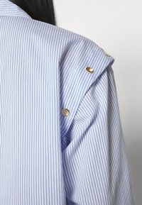 Chemise rayée bleu et blanc avec boutons-pression dorés à l'épaule, portée par une personne de dos sur un fond blanc uni.