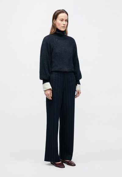 Mørk marineblå, blød high-neck sweater med ballonærmer kombineret med bredbenede bukser med nålestriber. Dybt burgunderrøde slip-on sko fremhæver looket.