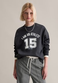 Czarna bluza z białym napisem "JESTEM SPORTOWCEM" i numerem "15" z przodu. Miękka tkanina, okrągły dekolt, ściągacze przy mankietach i u dołu. Szare spodnie.