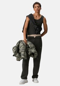 Schwarzes Tanktop, Tarnfleck-Steppjacke und schwarze Jogginghosen mit elastischem Bund. Sneakers und ein schwarzer Pullover locker über der Schulter getragen.