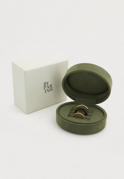 Βελούδινο πράσινο κουτί κοσμημάτων που περιέχει τρία δαχτυλίδια: δύο μεταλλικά με χρυσές και μαύρες επιφάνειες και ένα με υφή μαύρης επιφάνειας.