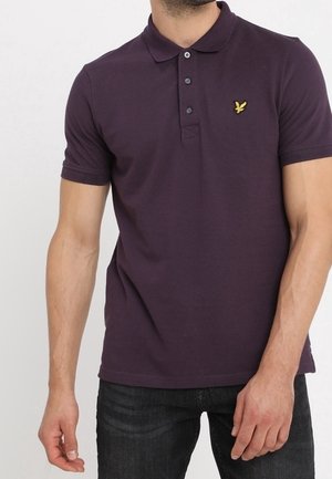 Mand iført en mørkelilla kortærmet polo med en gul fuglelogo på venstre bryst, kombineret med mørke jeans.