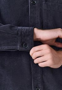 Dunkelbraunes Cordhemd mit vertikalen Rippungen, Knopfdetails an den Manschetten und einer Brusttasche. Der Stoff hat eine weiche, strukturierte Optik.