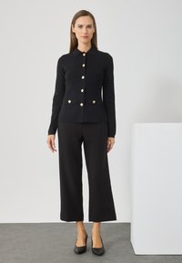 Svart knapp-up cardigan med gullknapper, parret med løse svarte bukser. Glatt tekstur og enkelt design, satt mot en nøytral bakgrunn.