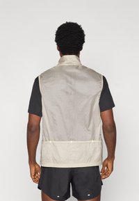 Ελαφρύ, μπεζ γιλέκο με ψηλό γιακά. Διαθέτει υφή υφάσματος, οπές αερισμού στο πίσω μέρος και ρυθμιζόμενο τελείωμα με κορδόνι. Το στυλ είναι μινιμαλιστικό και λειτουργικό.