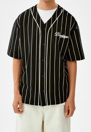 Mann trägt ein schwarzes kurzärmeliges Hemd mit weißen vertikalen Streifen und dem Schriftzug "Brooklyn", kombiniert mit weißen Hosen.