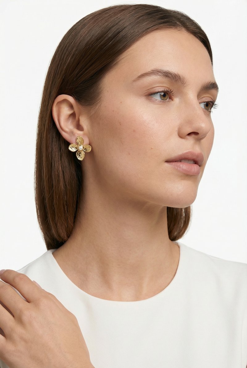 Mujer con cabello castaño liso lleva un pendiente dorado en forma de flor con una perla central, vestida con una blusa blanca, mirando hacia la derecha.