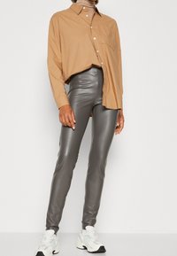 Oversized tan shirt met een zak, gecombineerd met strakke grijze neplederen broeken en witte sneakers met lichtblauwe accenten.