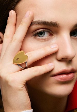 Primo piano di una persona che si tocca il volto, indossando un anello d'argento con un grande dettaglio dorato a forma di cuore sul dito medio.