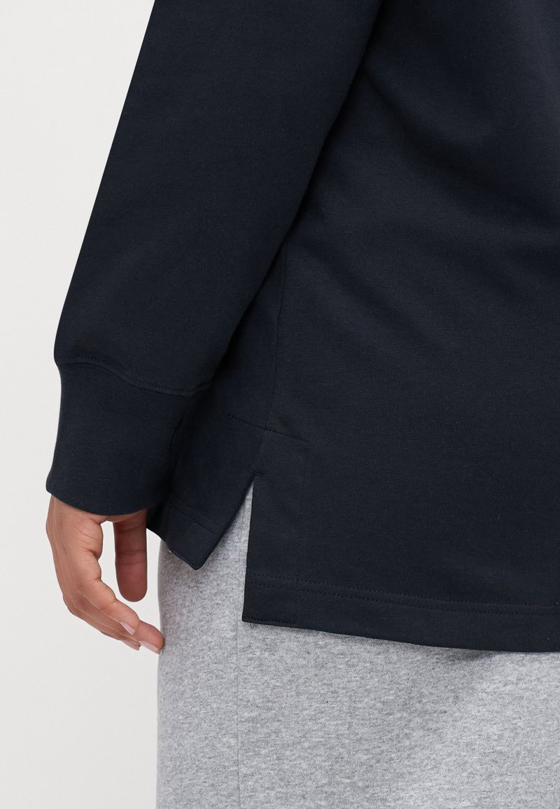Chemise à manches longues bleu marine avec col rond et fentes sur les côtés, réalisée en tissu coton lisse. Soulignée par une couture visible le long de la manche.