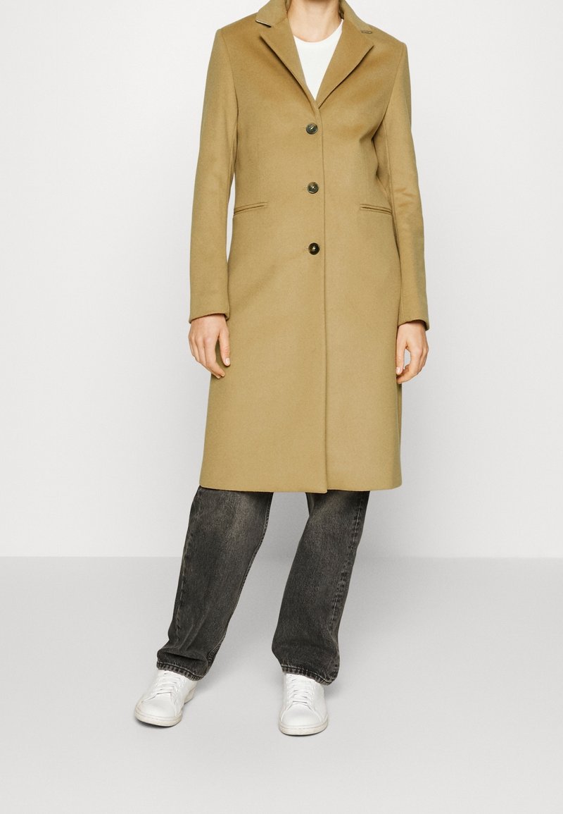 Cappotto di lana beige con collo a punta, tre bottoni frontali e tasche laterali, abbinato a jeans grigi e sneakers bianche.