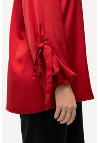 Blusa rossa con una texture morbida e liscia. Le maniche sono arricciate con un nastro e un elastico, creando un effetto morbido ai polsini.