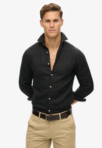 Camisa de lino negra con cuello abotonado, que presenta un corte holgado y mangas largas. Combinada con pantalones beige claros y un cinturón marrón.